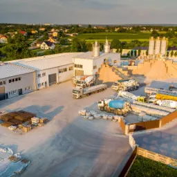 Widok z lotu ptaka na plac betoniarni: silosy, hałda piasku, betonomieszarki, ułożone prefabrykaty betonowe, zbrojenia i budynki w tle.