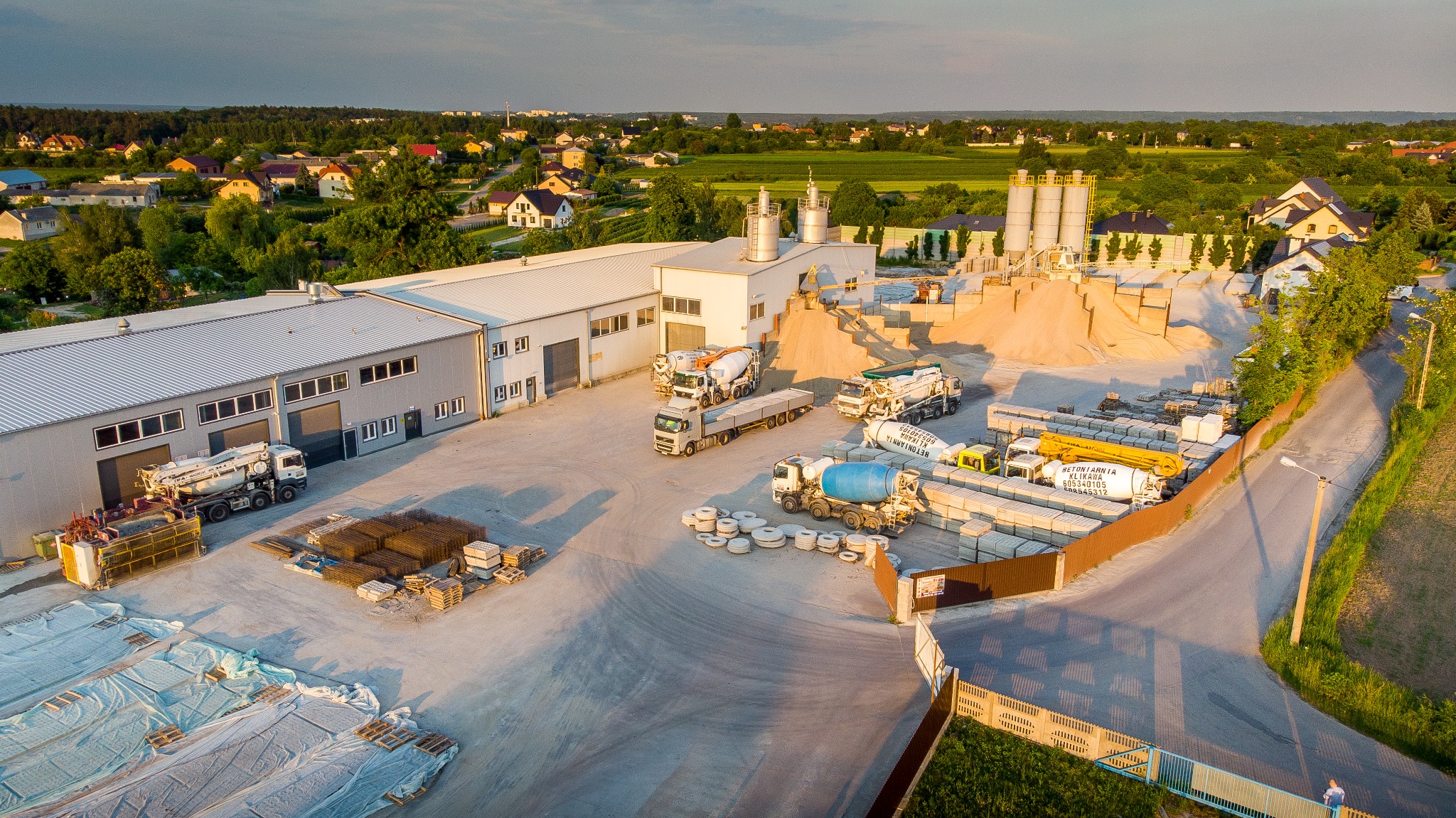 Widok z lotu ptaka na plac betoniarni: silosy, hałda piasku, betonomieszarki, ułożone prefabrykaty betonowe, zbrojenia i budynki w tle.