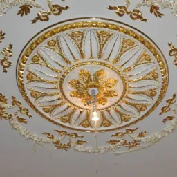 Dekoracyjny sufit ze sztukaterią w odcieniach bieli i złota, widok z dołu, w Zgierzu. Ozdobny wzór z motywami roślinnymi i centralnym elementem przypominającym kwiat.