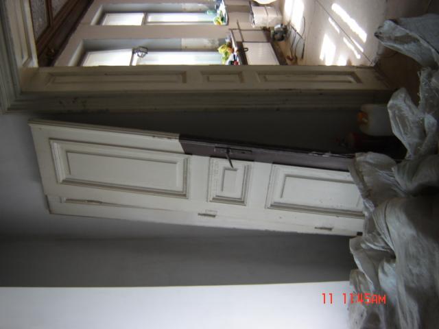 Stara, biała, częściowo pomalowana rama drzwiowa z widocznymi uszkodzeniami i workami z gruzem w tle, sugerująca etap remontu lub renowacji.