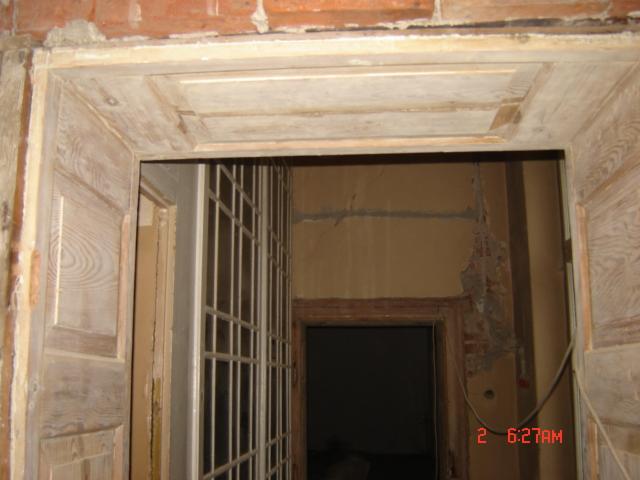 Drewniana, odnowiona rama drzwiowa z dekoracyjnymi panelami, widoczna na tle wnętrza w trakcie prac wykończeniowych, z odsłoniętą cegłą i elementami instalacji.