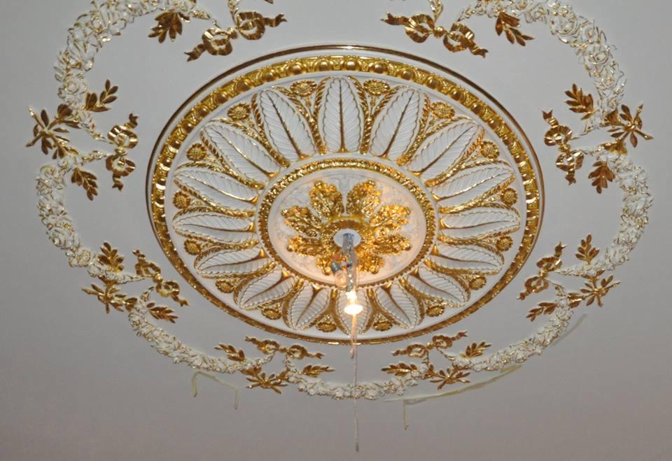 Dekoracyjny sufit ze sztukaterią w odcieniach bieli i złota, widok z dołu, w Zgierzu. Ozdobny wzór z motywami roślinnymi i centralnym elementem przypominającym kwiat.