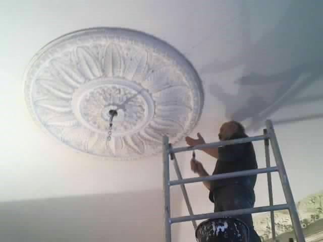 Malowanie sufitu: mężczyzna na drabinie odświeża dekoracyjny, okrągły element sztukaterii pędzlem. Widoczny kubeł z farbą. Praca na wysokości.