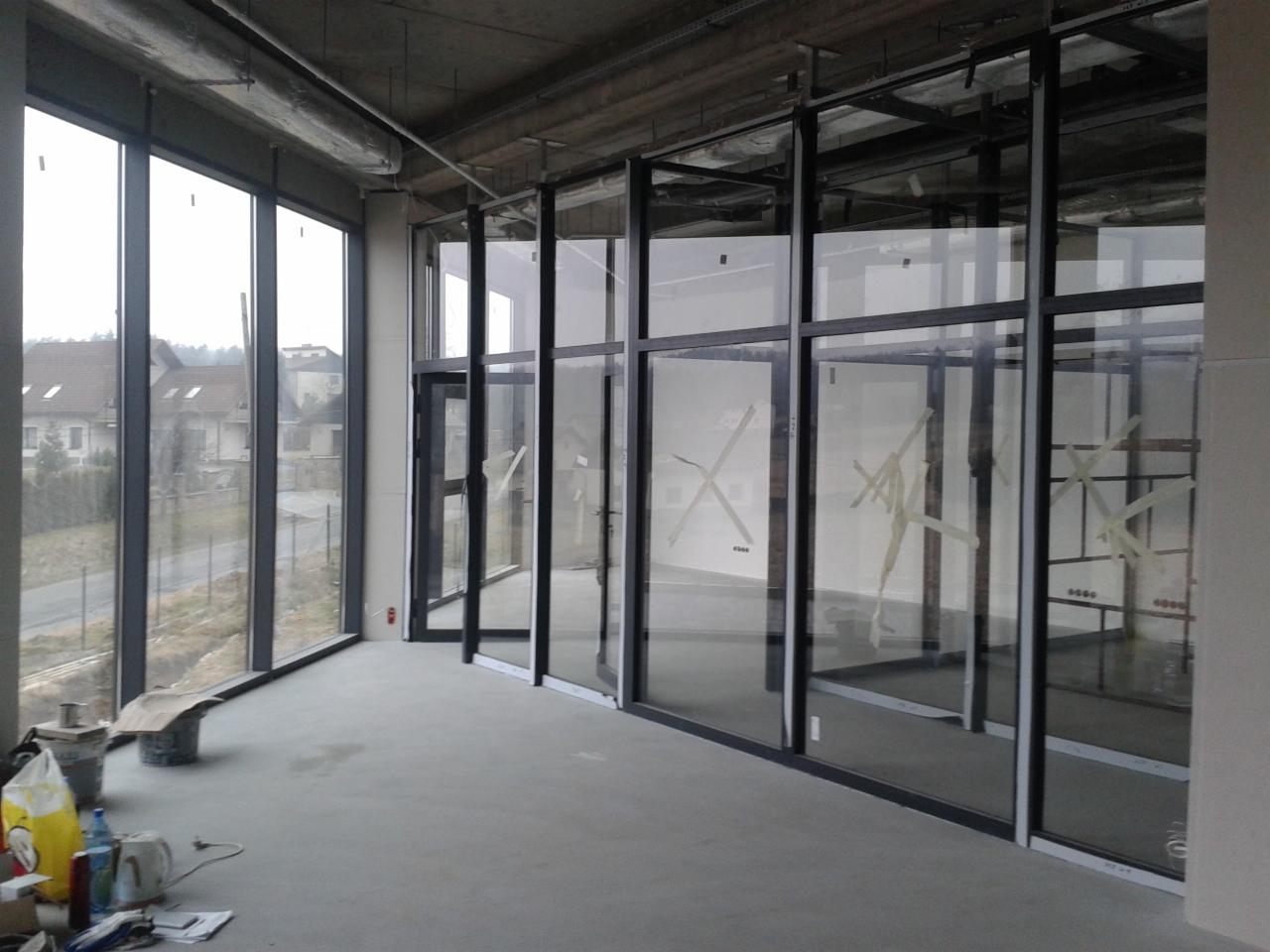Wnętrze hali w budowie z zamontowanymi oknami i szklanymi ścianami działowymi, zabezpieczonymi taśmą. Widok na zewnątrz przez duże okna.