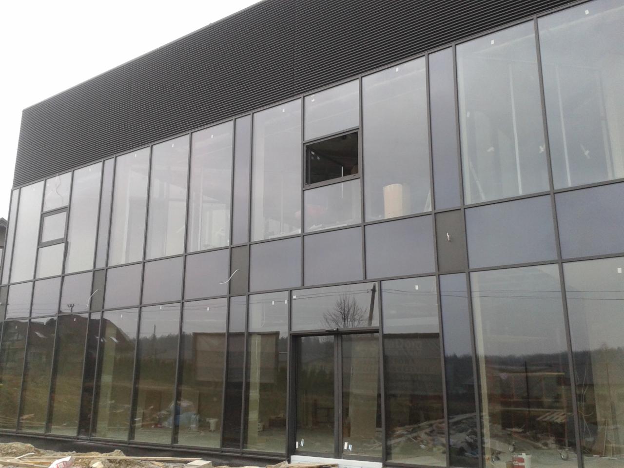 Nowoczesna hala z szarymi płytami warstwowymi i dużymi oknami. Część okien jest uchylona. Widoczne materiały budowlane na zewnątrz.