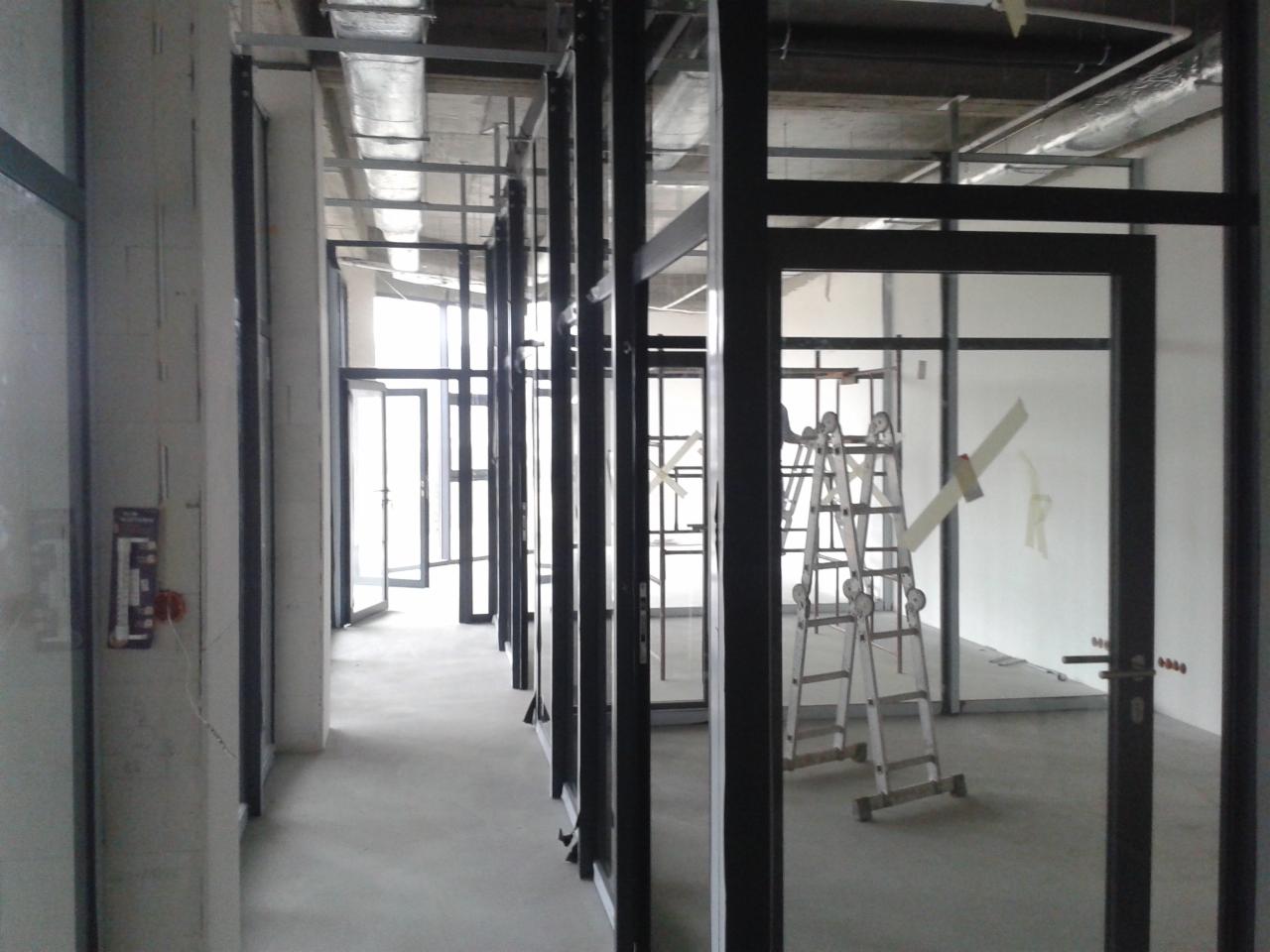 Wnętrze biurowe w trakcie tworzenia, widoczne metalowe ramy szklanych ścian działowych i drzwi, drabina aluminiowa w tle, surowy betonowy sufit z instalacją wentylacyjną, widok z perspektywy...