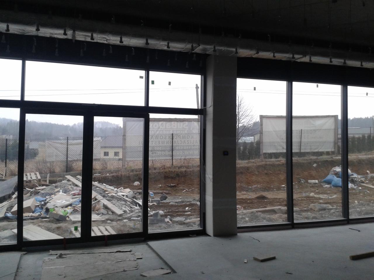 Czarne, panoramiczne okna w surowym stanie, z widokiem na teren budowy i częściowo odsłonięte reklamy. Wnętrze w trakcie wykończenia, surowy beton.