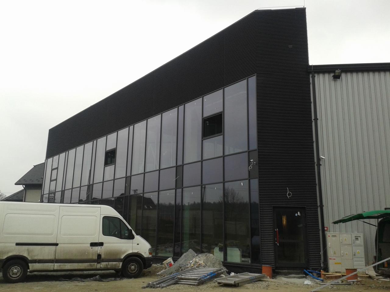 Nowoczesna hala z czarną elewacją i dużymi oknami, częściowo ukończona. Na pierwszym planie gruz i zaparkowany biały bus.