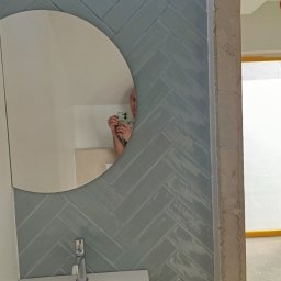 Daros Arkadiusz - Łazienka po remoncie: błękitne płytki ułożone w jodełkę, okrągłe lustro z odbiciem osoby robiącej zdjęcie, umywalka z szafką. Widoczny surowy fragment ściany.