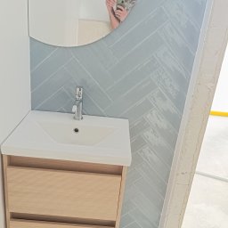 Daros Arkadiusz - Nowoczesna łazienka z umywalką na szafce z jasnego drewna i lustrem. Ściana wyłożona błękitnymi płytkami w jodełkę. W lustrze odbija się osoba robiąca zdjęcie.