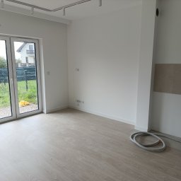 Daros Arkadiusz - Jasne wnętrze po remoncie: świeżo pomalowane ściany, panele podłogowe imitujące drewno, okno z widokiem na ogród. Częściowo widoczna drabina i rura wentylacyjna.