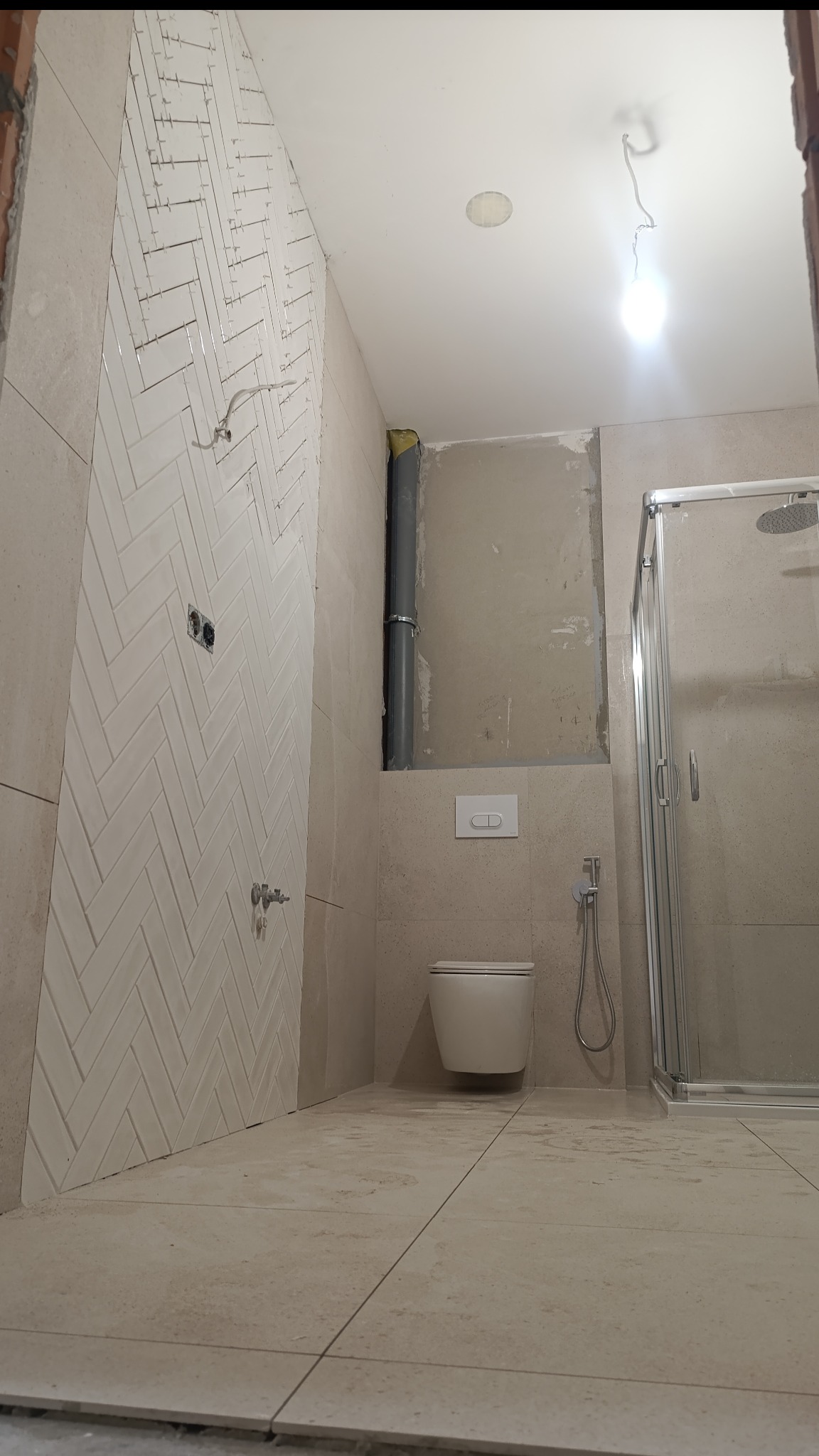 Łazienka w trakcie remontu: ściana z białymi płytkami ułożonymi w jodełkę, widoczna toaleta, kabina prysznicowa i fragment instalacji. Prace wykończeniowe w toku.