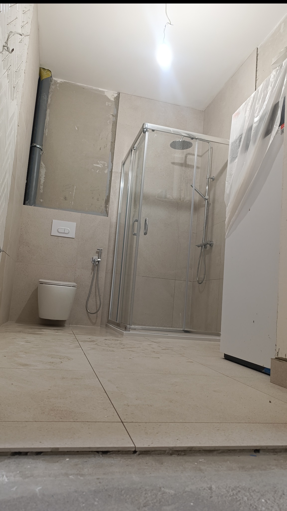 Wnętrze łazienki w trakcie remontu: kabina prysznicowa, toaleta podwieszana, beżowe płytki na ścianach i podłodze. Widoczne niedokończone prace wykończeniowe.