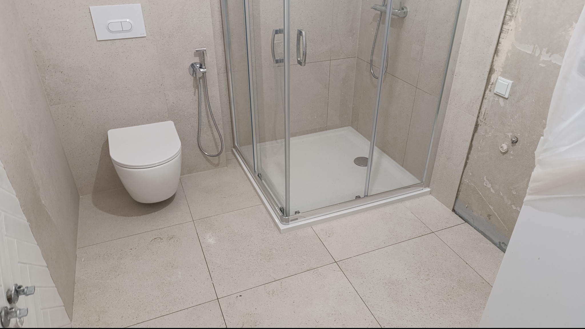 Jasna łazienka po remoncie: kabina prysznicowa z brodzikiem, podwieszana toaleta z bidetką, beżowe płytki podłogowe i ścienne. Fragment ściany w trakcie wykończenia.