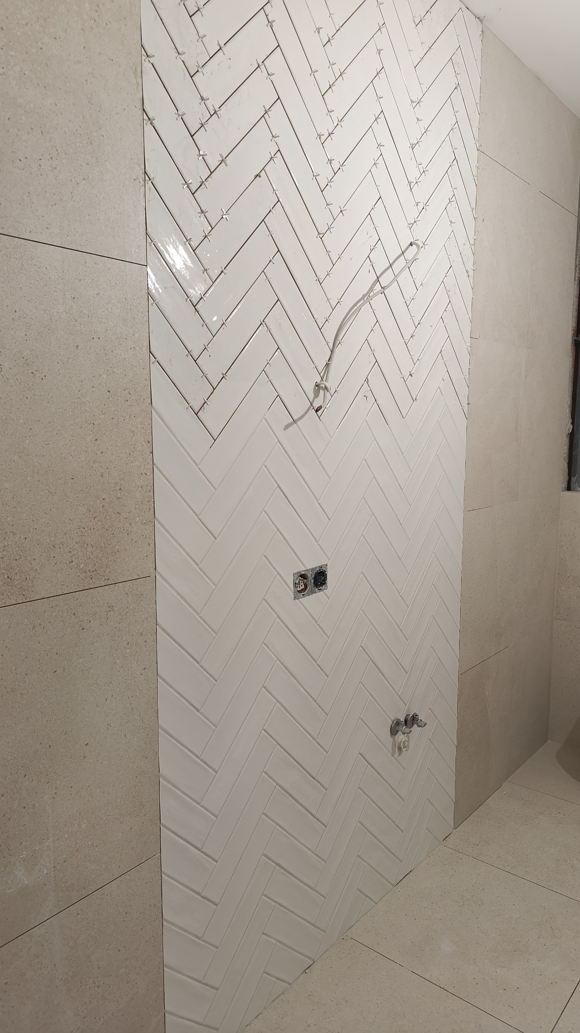 Ściana w trakcie wykańczania z białymi, błyszczącymi płytkami ułożonymi w jodełkę. Widoczne krzyżyki dystansowe, gniazdka elektryczne i wyprowadzenie instalacji wodnej.