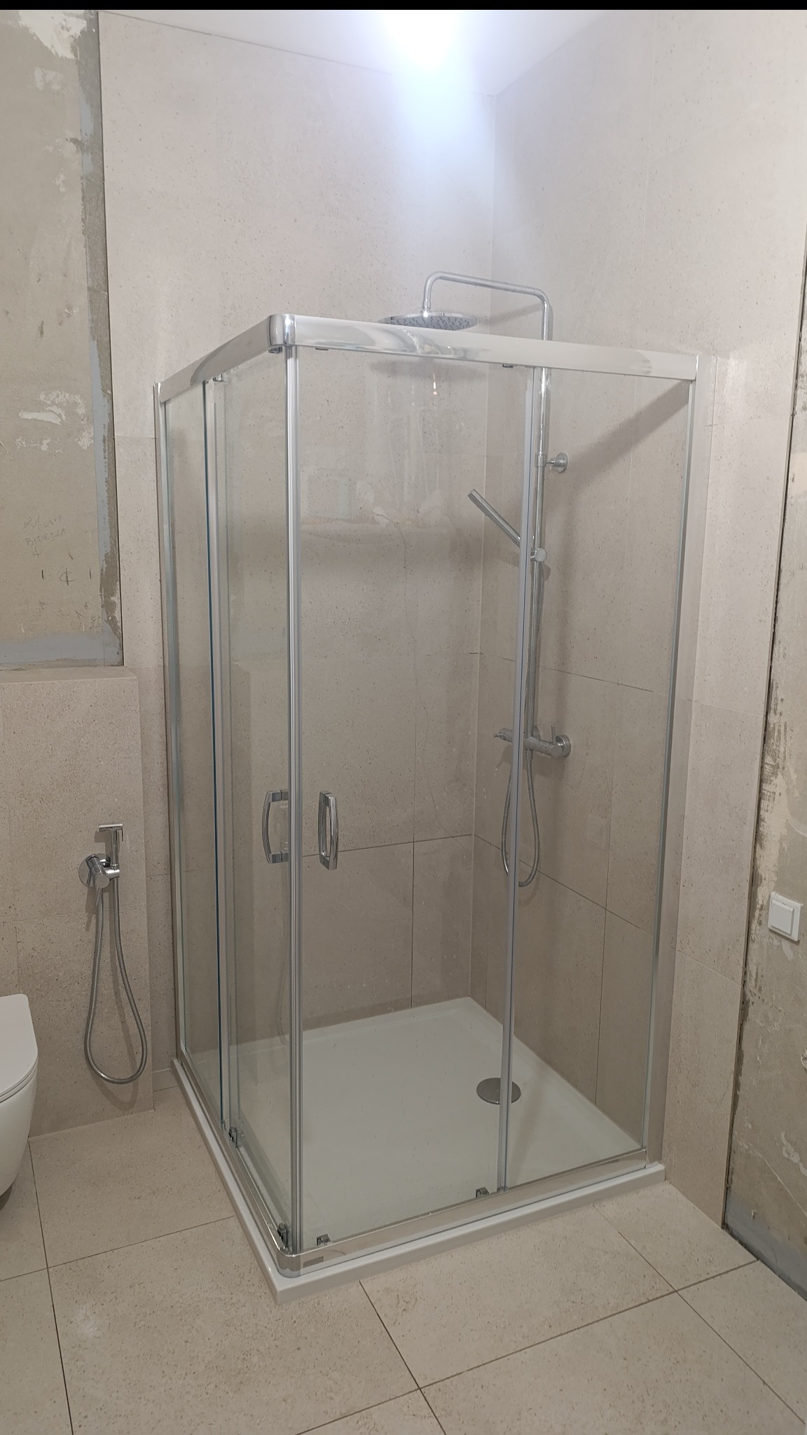 Nowoczesna kabina prysznicowa z chromowanymi profilami i deszczownicą, zamontowana w łazience z beżowymi płytkami. Widoczne elementy instalacji wodnej i fragment toalety.