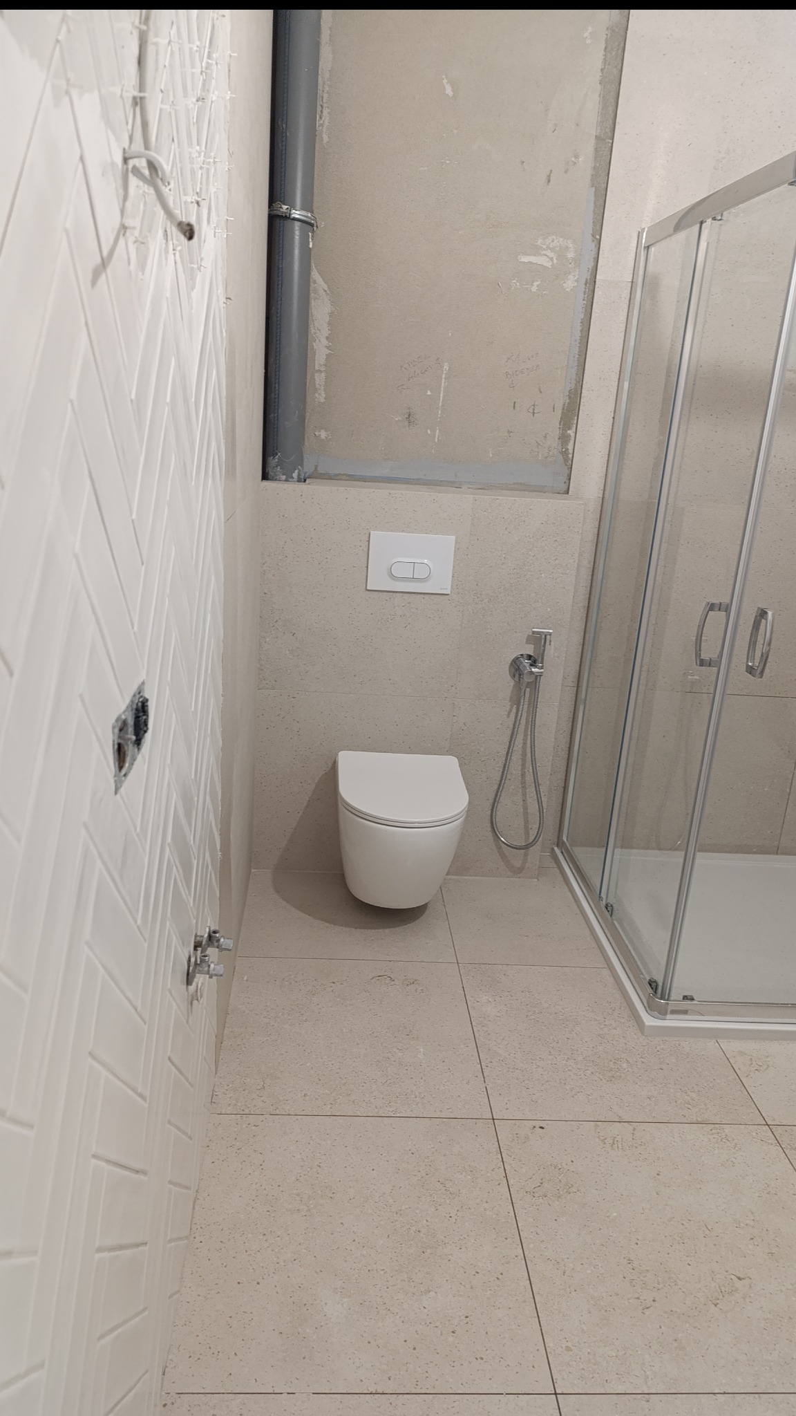 Jasna łazienka w trakcie remontu: biała ściana z ułożoną glazurą w jodełkę, podtynkowa toaleta, bidetka i kabina prysznicowa. Widoczne niedokończone prace.