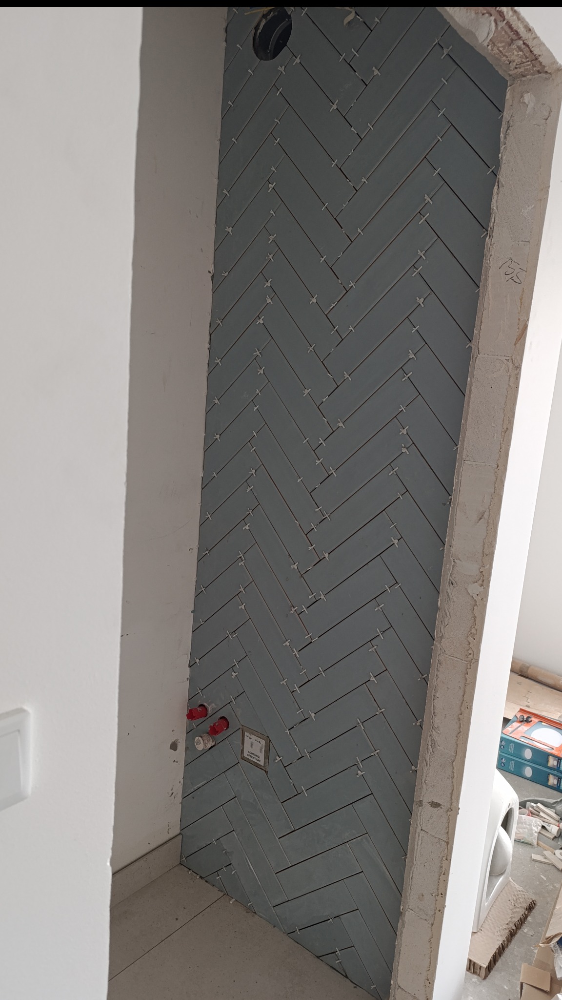 Ściana wyłożona szarymi, podłużnymi płytkami ułożonymi w jodełkę, z widocznymi krzyżykami dystansowymi i fragmentami instalacji wodnej. Surowy stan wykończenia.