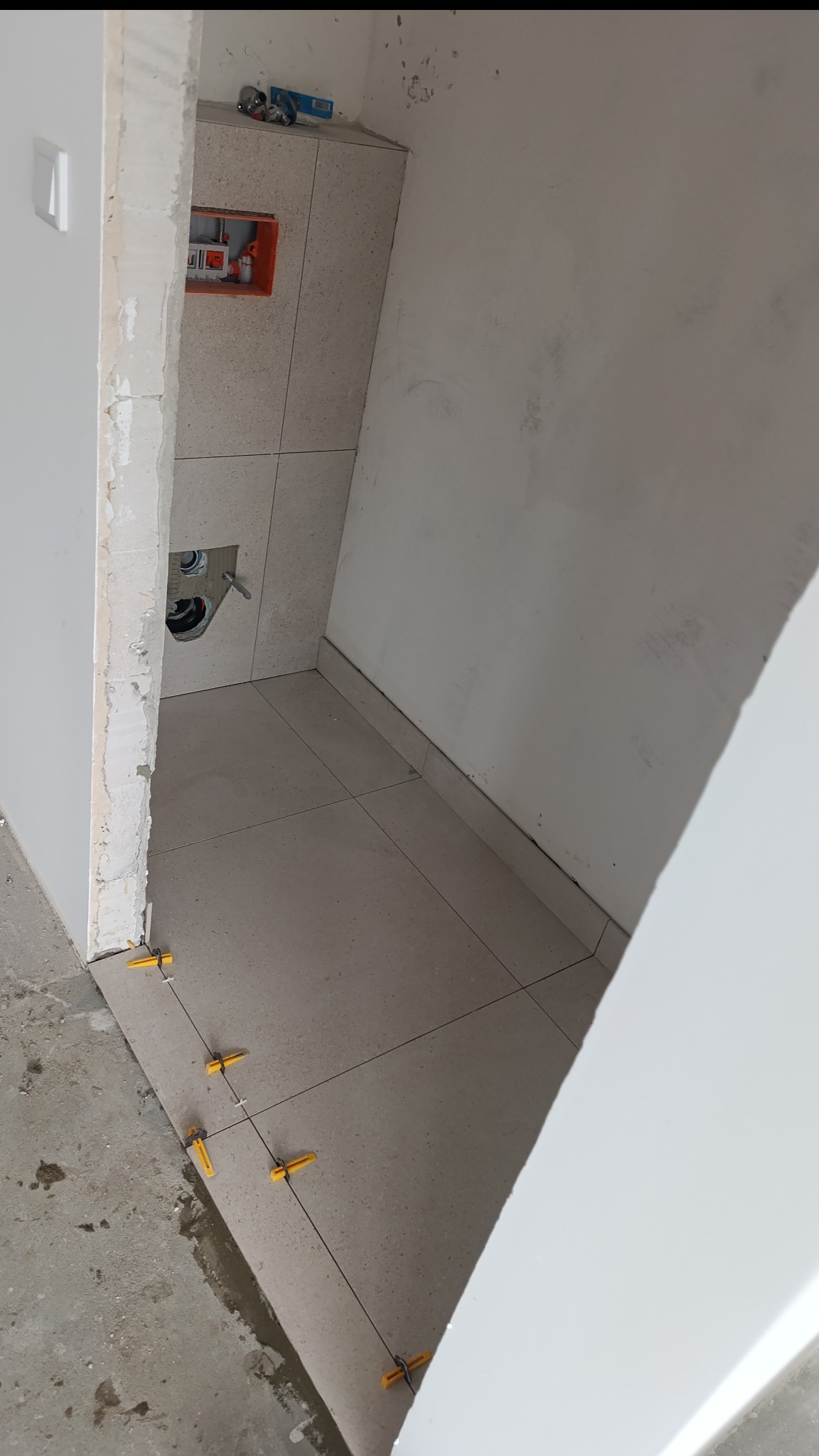 Wnętrze łazienki w trakcie remontu. Widoczne ułożone płytki podłogowe z żółtymi krzyżykami dystansowymi oraz fragment ściany z instalacją hydrauliczną.