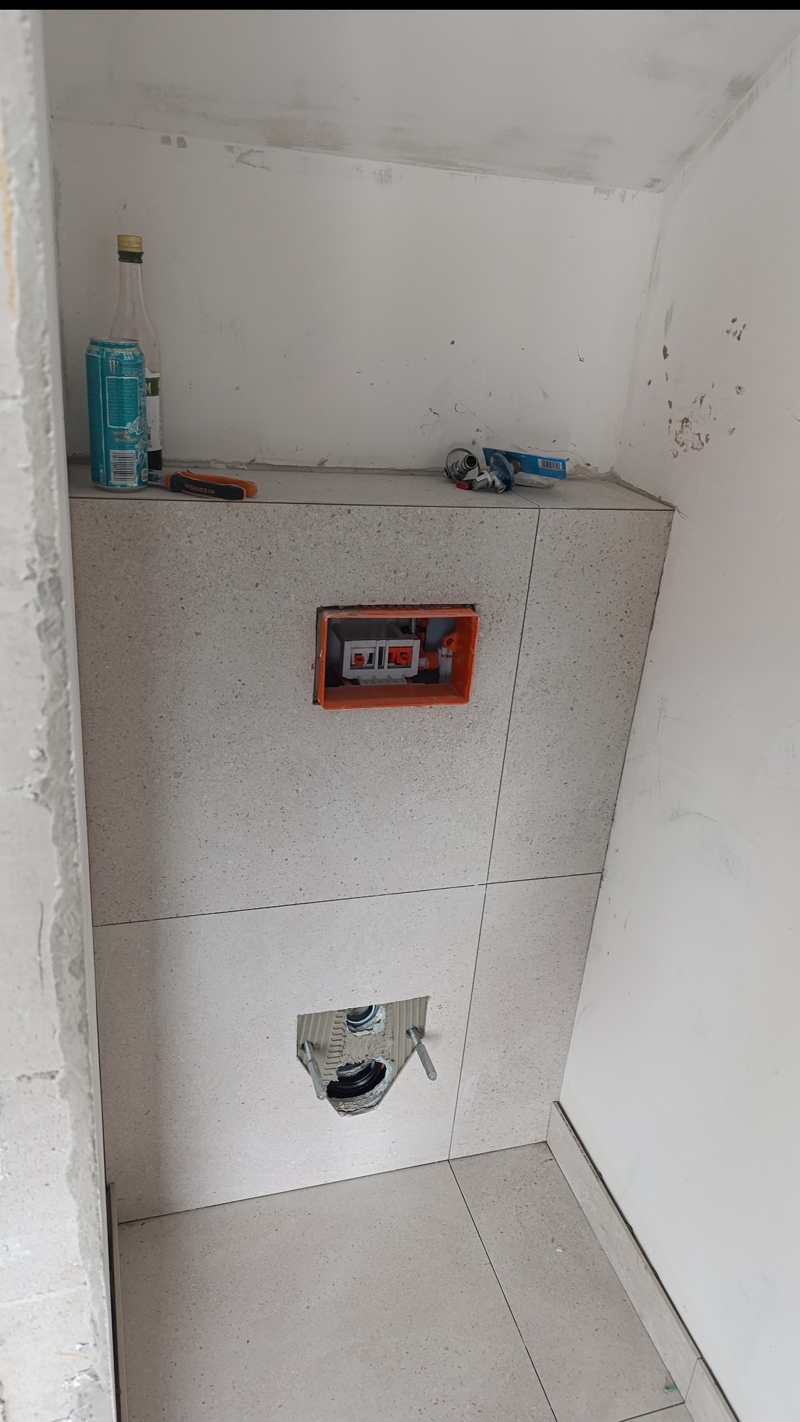 Ściana w trakcie wykańczania z zamontowaną instalacją podtynkową WC, obłożona jasnymi płytkami imitującymi kamień. Widoczne narzędzia i puszka z instalacją.