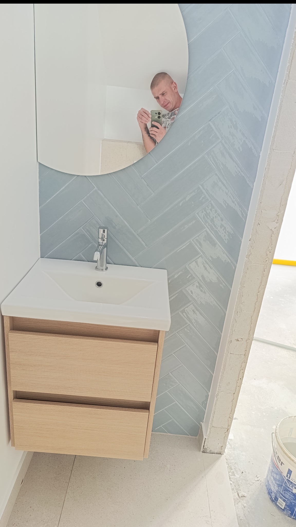 Nowoczesna łazienka z umywalką na szafce z jasnego drewna i lustrem. Ściana wyłożona błękitnymi płytkami w jodełkę. W lustrze odbija się osoba robiąca zdjęcie.