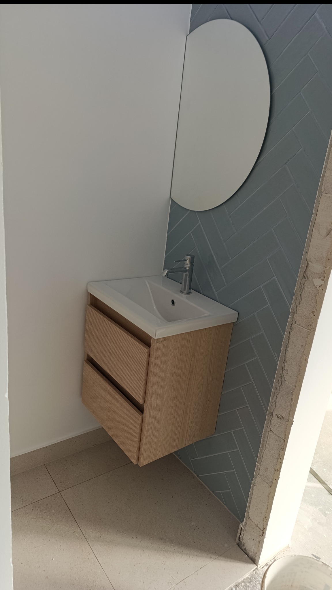 Nowoczesna łazienka z umywalką nablatową na szafce z drewnianymi frontami, okrągłe lustro i szare płytki ułożone w jodełkę na ścianie.