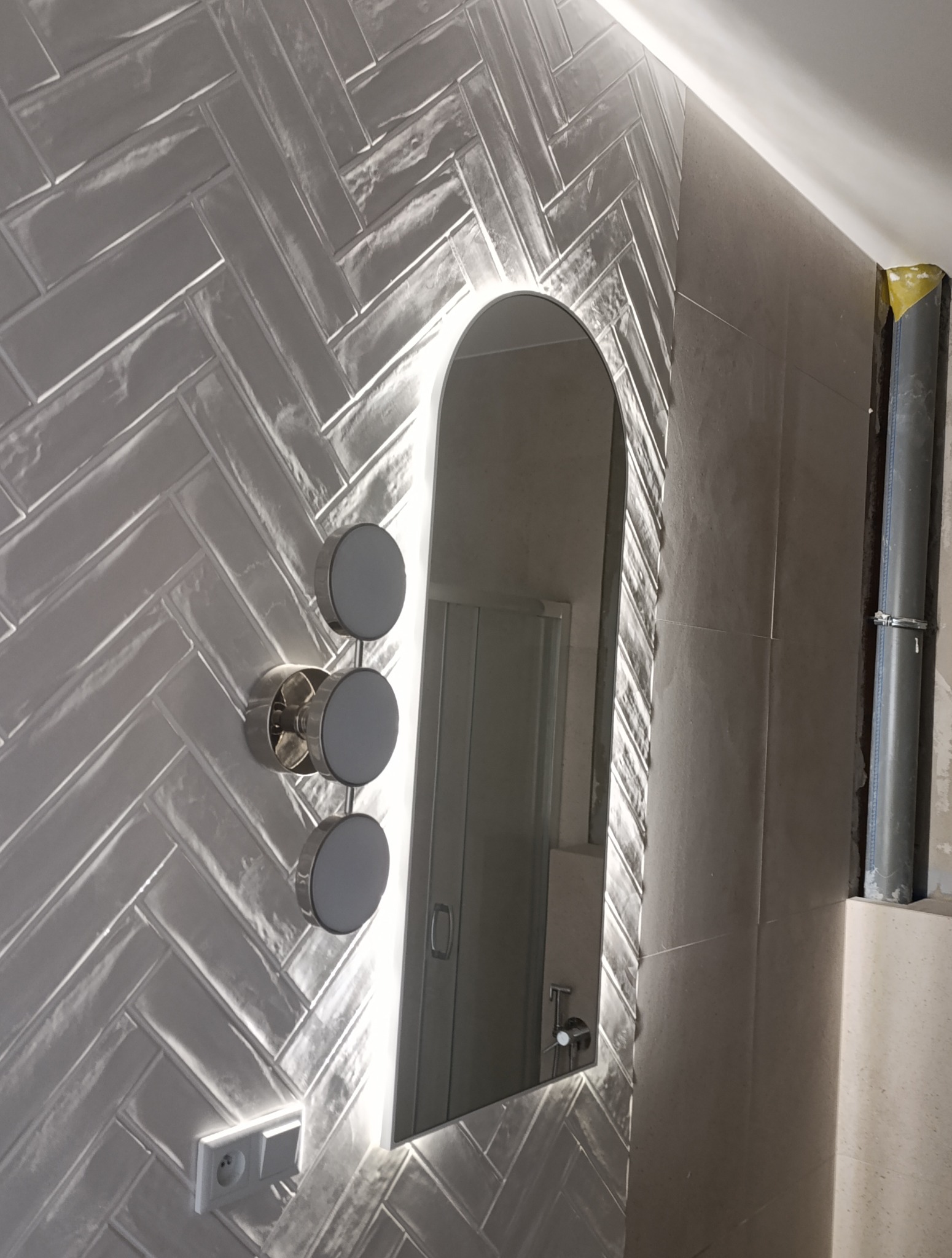 Biała łazienka z lustrem podświetlanym LED i płytkami ułożonymi w jodełkę. Widoczny fragment instalacji. Nowoczesny design z elementami w trakcie wykończenia.