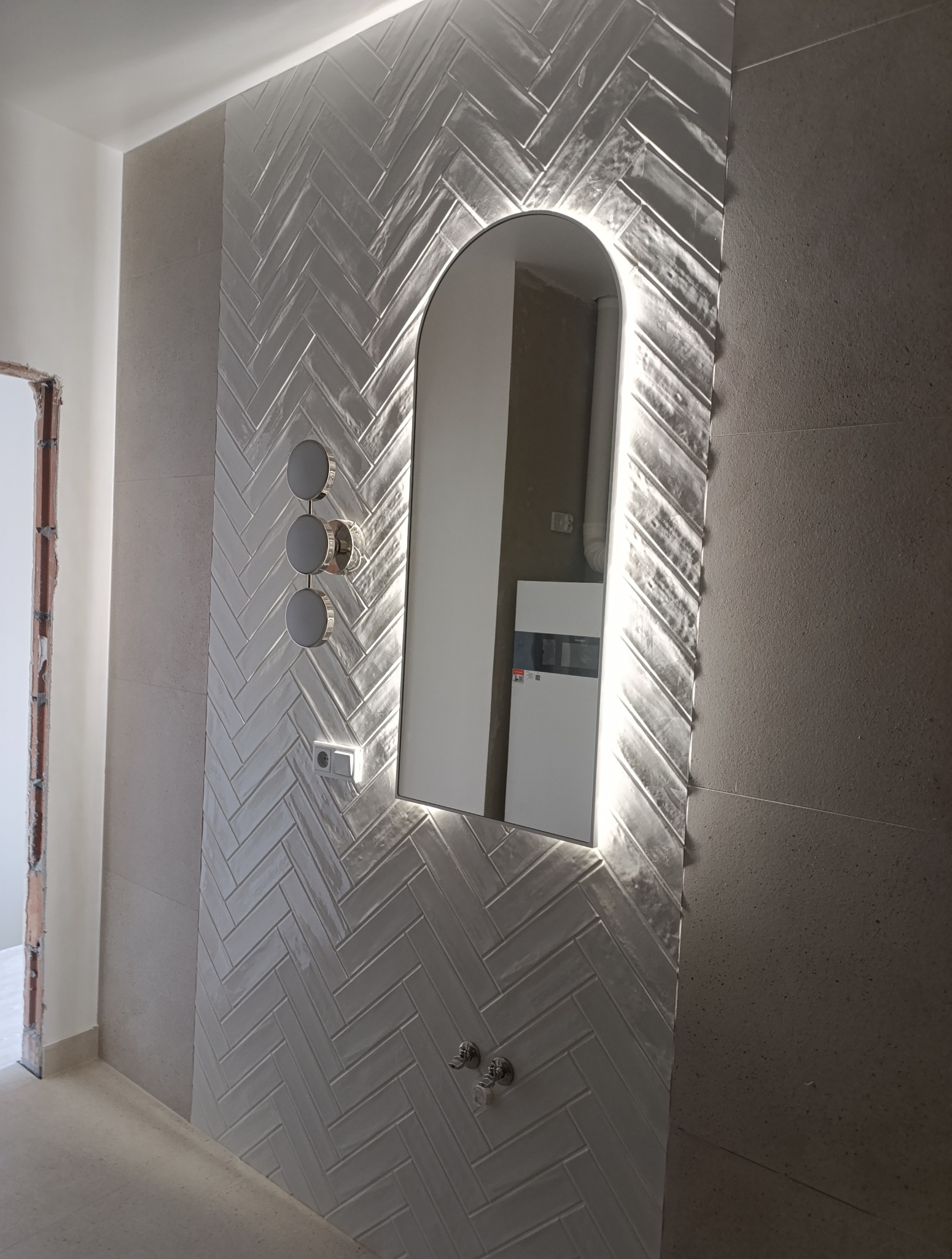 Wykończona ściana w łazience z ułożonymi w jodełkę białymi płytkami, podświetlane lustro w kształcie łuku, bateria i włącznik światła. Widoczny fragment niedokończonego wejścia.