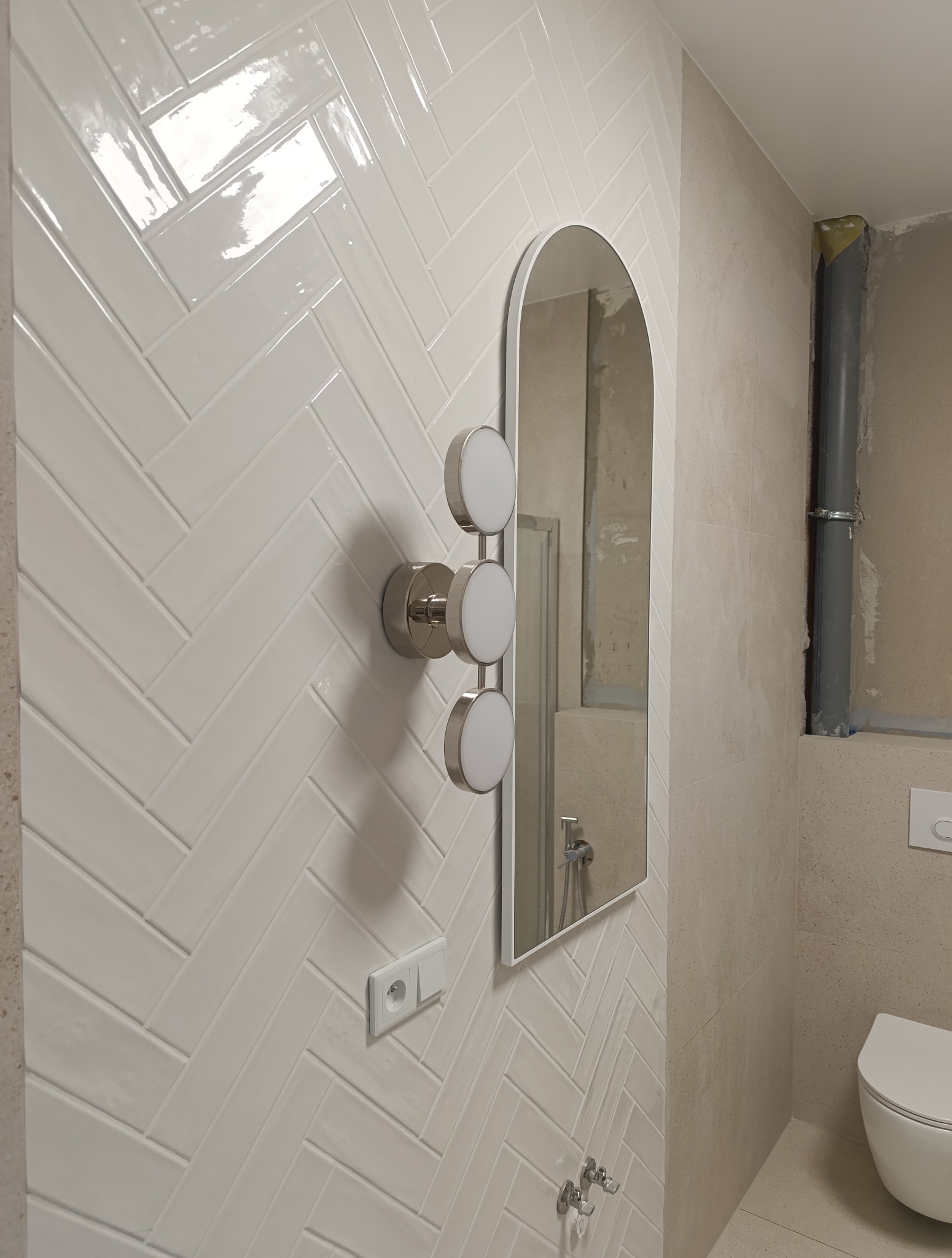 Wykończona łazienka z białymi, błyszczącymi płytkami ułożonymi w jodełkę, nowoczesnym oświetleniem i owalnym lustrem. Widoczna toaleta i fragment instalacji.