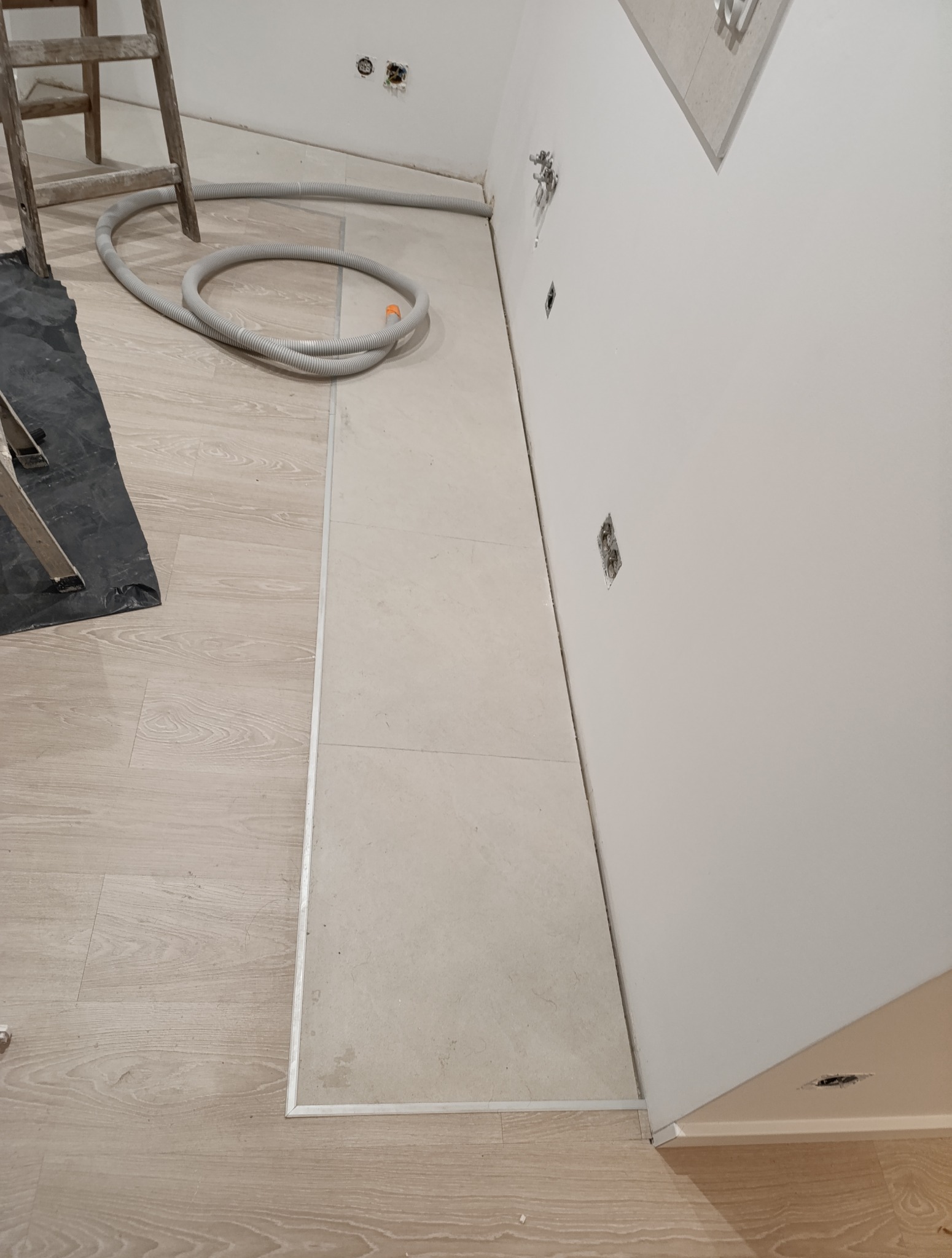 Fragment podłogi w trakcie remontu: ułożone jasne płytki z metalową listwą oddzielającą od paneli, drabina i rura wentylacyjna w tle, gniazdka elektryczne na ścianie.