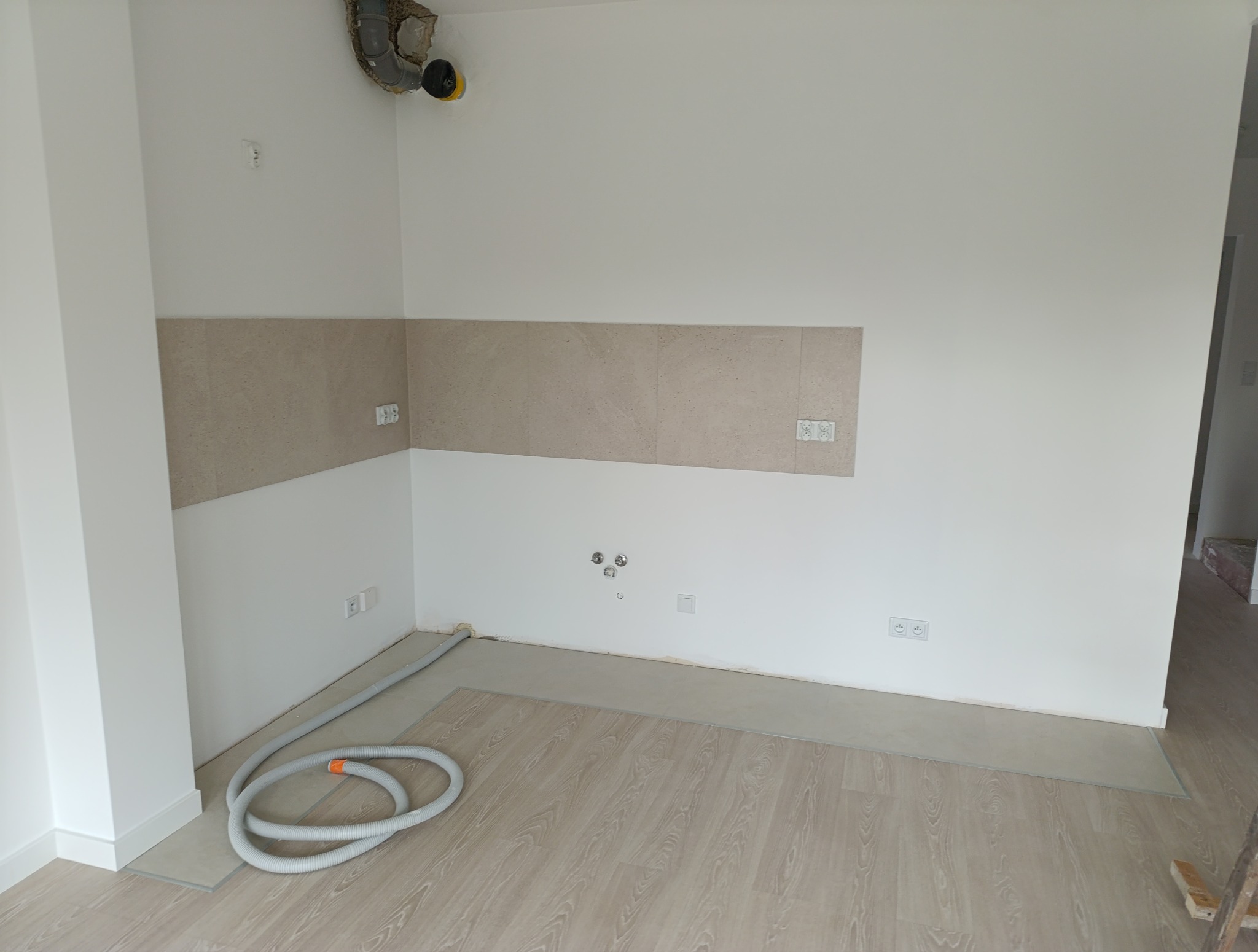 Wnętrze kuchni w trakcie remontu: położone panele podłogowe, częściowo wykończona ściana z beżowymi płytkami, widoczne instalacje elektryczne i rura wentylacyjna.