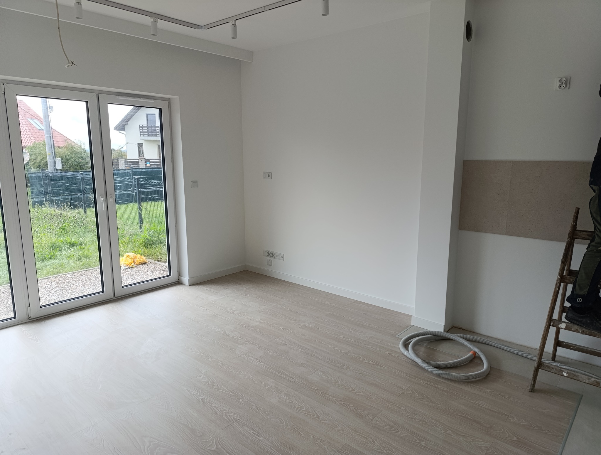 Jasne wnętrze po remoncie: świeżo pomalowane ściany, panele podłogowe imitujące drewno, okno z widokiem na ogród. Częściowo widoczna drabina i rura wentylacyjna.