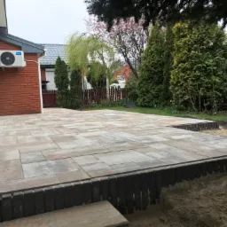 Nowoczesny taras z szarej kostki brukowej ułożonej na posesji z widokiem na trawnik, drzewa i dom z klimatyzatorem na ścianie.