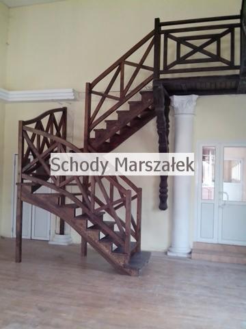 Dwubiegowe schody drewniane z balustradą o dekoracyjnych krzyżowych wzorach, wsparte na kolumnie. Widok na jasne wnętrze z podłogą imitującą drewno.