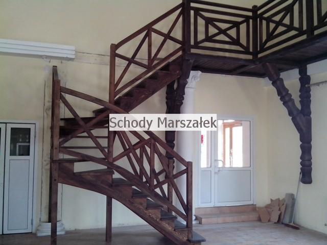 Kreatywne schody drewniane z balustradą krzyżową, prowadzące na antresolę z ozdobnymi kolumnami. Ciemne drewno kontrastuje z jasnymi ścianami i drzwiami.