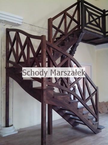 Ciemnobrązowe, drewniane schody z balustradą o ażurowym wzorze krzyżowym, prowadzące na antresolę. Widoczne detale wykończenia i konstrukcja schodów.