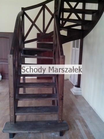 Kręte, ciemne schody drewniane z ażurową balustradą w jasnym wnętrzu. Widoczny fragment podłogi i drzwi w tle. Ujęcie z dołu.