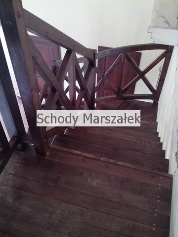 Ciemne, drewniane schody z ozdobną balustradą w stylu rustykalnym. Widok z góry na stopnie i fragment ściany z białym wykończeniem. Charakterystyczny wzór balustrady.