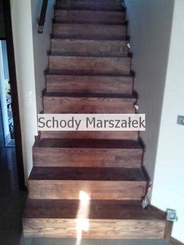 Drewniane schody wewnątrz domu, z widocznym usłojeniem drewna i detalami wykończenia, patrząc od dołu ku górze. Widoczne mocowania poręczy.