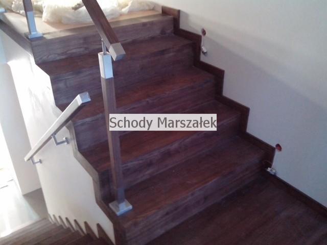 Schody drewniane z metalową balustradą, widok z dołu. Stopnie w odcieniu ciemnego brązu, z wyraźnym usłojeniem drewna. Balustrada z elementami srebrnymi i drewnianą poręczą.