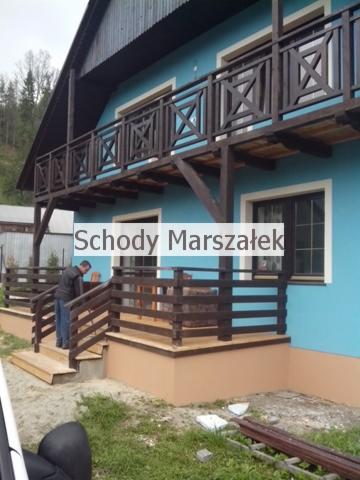 Błękitny dom z drewnianą balustradą balkonową o wzorze krzyży, dopasowaną do drewnianych schodów z poręczą. Osoba wchodzi po schodach.