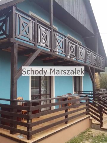 Drewniana balustrada balkonowa z geometrycznym wzorem krzyży, ciemnobrązowa, na tle błękitnej elewacji domu. Widok z dołu, częściowo zasłonięte krzesła i stoliki.