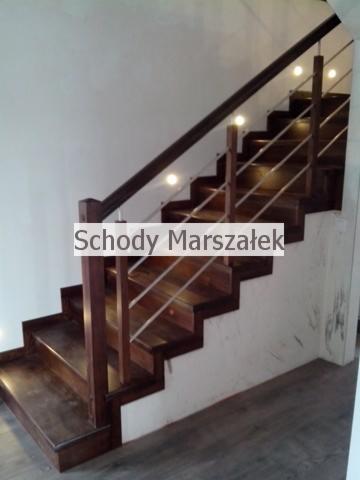 Drewniane schody z podświetleniem LED i metalową balustradą, widok z dołu, ukazujące solidne wykonanie i nowoczesny design. Ciemne drewno kontrastuje z bielą ścian.