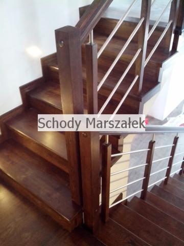 Ciemne, drewniane schody z balustradą z metalowych prętów, widziane z góry. Charakterystyczne, kwadratowe słupki balustrady z widocznymi śrubami.