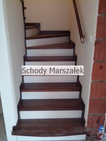 Nowoczesne schody z ciemnego drewna i białych podstopnic, z widoczną poręczą. Elementy wykończeniowe w kolorze miedzianym. Widok z dołu na wprost.