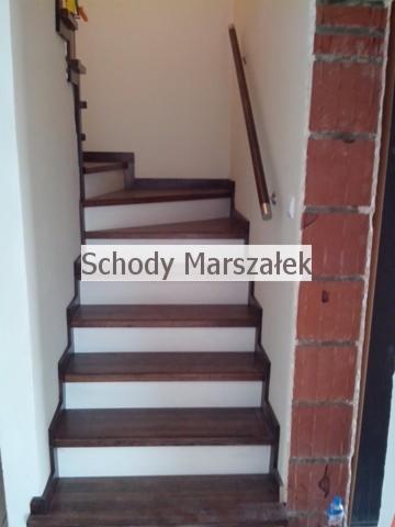 Drewniane schody z białymi podstopnicami i ciemnymi stopniami. Widoczna drewniana poręcz przy ścianie i fragment ceglanej obudowy.