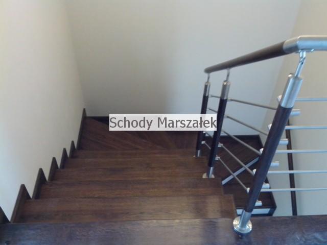 Kątowe ujęcie schodów z ciemnego drewna z metalową balustradą i drewnianym poręczem. Widok z góry na stopnie i fragment białej ściany w tle.