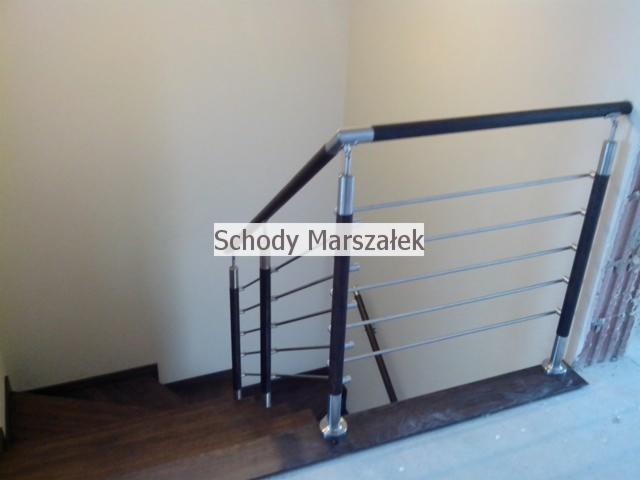Fragment schodów z balustradą: drewniane stopnie, metalowe słupki w czarnym kolorze i poziome, stalowe rurki. Minimalistyczny design, jasne tło ściany.