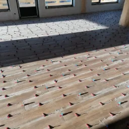 Brązowe płytki podłogowe imitujące drewno, ułożone z systemem poziomowania płytek (czerwone i niebieskie klipsy) w jasnym, surowym wnętrzu z drewnianym słupem i oknami.