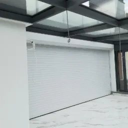 Biała roleta zewnętrzna zamontowana w przeszklonym pomieszczeniu, widoczne przewody instalacyjne i marmurowa podłoga.